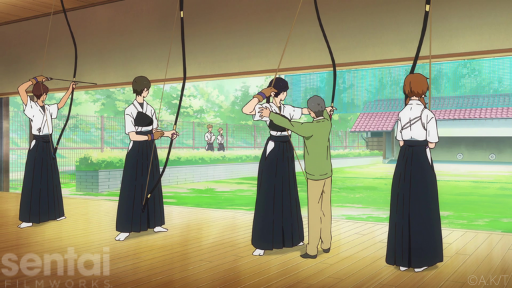 A row of young women aim kyudo bows at a row of distant targets while their teacher corrects their shooting form.