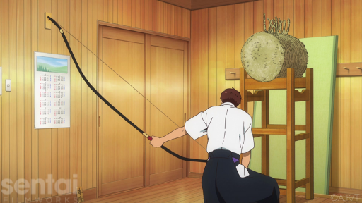 A character from the Tsurune anime crouches with his back to the viewer. He holds a long kyudo bow, showing its asymmetrical structure.