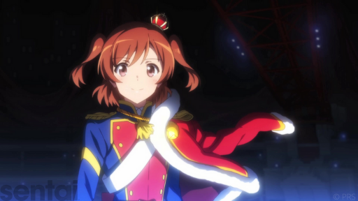 Karen of Revue Starlight gazes happily toward the viewer. She has brown eyes, brown pigtails and wears a small crown. Her outfit comprises a blue and red military uniform with a red shoulder-cape.