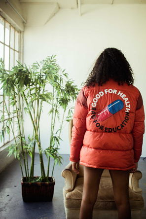 A model with curly brown hair stands with her back to the camera. She wears a red puffer jacket that looks like the jacket from the movie Akira, which features a blue and white pill and the words "GOOD FOR HEALTH" and "BAD FOR EDUCATION."